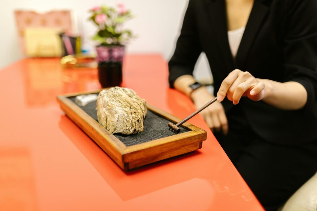 A woman in a black suit rakes a mini Zen garden on a red desk, symbolizing relaxation and mindfulness.